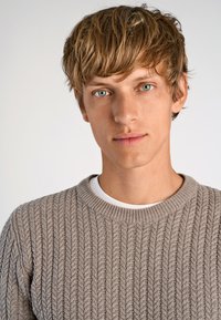 Ljusbeige tröja med ribbad textur och rund hals, bärs över en vit T-shirt. Modellen har kort, rufsigt hår och blå ögon.