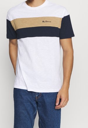 Homme portant un T-shirt blanc à manches courtes avec des rayures horizontales beige et marine et un logo brodé "Ben Sherman", associé à un jean bleu.