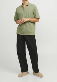Světlá zelená polo košile se zipovým límcem, kombinovaná s volnými černými kalhotami a světlými slip-on botami, vykazující uvolněný střih a hladkou strukturu.