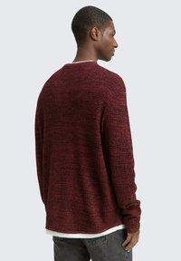 Man in een gestructureerde donkerrode trui met lange mouwen en donkergrijze jeans, gezien van achteren tegen een effen lichte achtergrond.