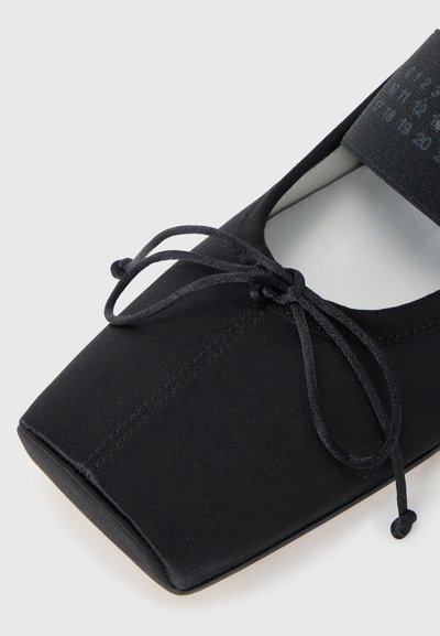 Black ballet flat with a square toe, fabric upper, thin laces, and a cushioned insole. Features a stitched seam design and elastic strap.
