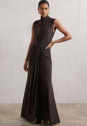 Kobieta ubrana w czarną, bezrękawową suknię z wysokim kołnierzem, sięgającą do podłogi, z drapowanymi detalami, stojąca na tle gładkiej, jasno szarej ściany.