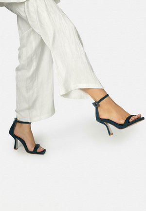 Piernas con pantalones blancos de pierna ancha, calzando sandalias de tacón alto azul marino con tiras en el tobillo y hebilla decorativa.