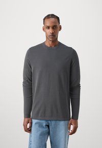 Cămașă din bumbac gri cu mâneci lungi și gât rotund. Țesătura este netedă și ajustată, combinată cu blugi de denim albaștri deschiși.