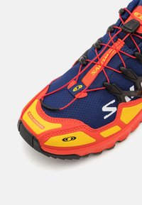 Zapatilla de correr en azul y rojo con parte superior de malla, detalles en negro, suela acolchada y un distintivo acento amarillo en el lateral.