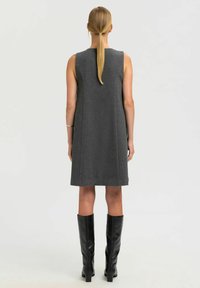 Ärmelloses graues Kleid aus einem Wollmischgewebe, knielang, mit gerader Silhouette. Trägt dazu hohe schwarze Lederstiefel.