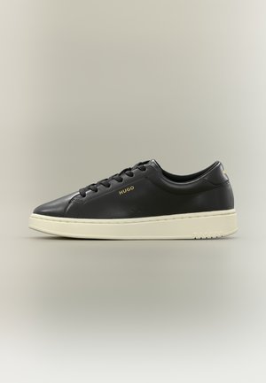 Basket basse noire avec lacets noirs, semelle blanche et logo doré "HUGO" sur le côté et le talon sur un fond uni.