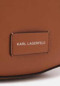 Bolso de cuero marrón con superficie texturizada y costura en contraste. Presenta una etiqueta rectangular grabada con "KARL LAGERFELD" en blanco.