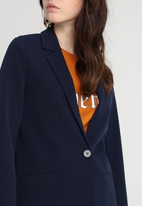 Femme portant un blazer bleu marine sur une chemise orange avec un texte blanc partiel, montrée des épaules au milieu du torse sur un fond uni.