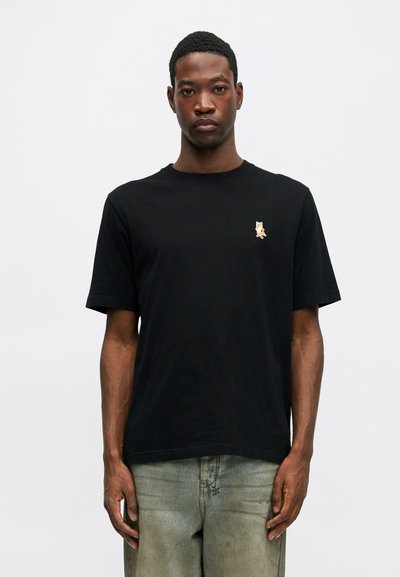 Hombre con una camiseta de manga corta negra con un pequeño parche de un zorro naranja en el lado izquierdo del pecho y jeans claros desgastados, de pie frente a un fondo blanco.