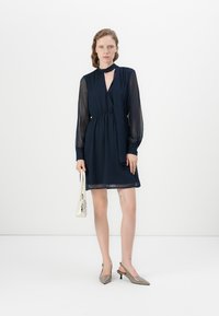 Μαρινί μπλε φόρεμα με μακριά διάφανα μανίκια, V ντεκολτέ και ζωνάκι στη μέση. Το μοντέλο κρατάει μια άσπρη τσάντα, συνδυασμένη με γκρίζα μυτερά παπούτσια.