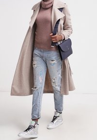 Persona con un abrigo largo beige, suéter de cuello alto marrón, jeans rasgados manchados de pintura, zapatillas altas iridiscentes y llevando un bolso cruzado azul marino.