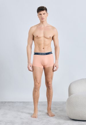 Uomo in piedi a piedi nudi che indossa boxer Lacoste color pesca, con sfondo chiaro uniforme e una poltrona bianca con trama vicina.