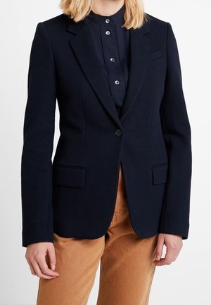 Femme portant un blazer noir ajusté sur une chemise noire à boutons et un pantalon brun clair, debout devant un fond blanc uni.