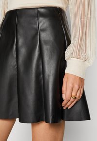 Schwarzer Lederrock mit ausgestelltem Design und Falten. Reißverschluss an der Seite. Kombiniert mit einem transparenten, gerippten Langarmshirt und einem goldenen Ring.