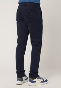 Chinos blu navy con vestibilità affusolata, dotati di due tasche posteriori con chiusura a bottone. Indossati con sneakers bianche e blu, mostrando un tessuto testurizzato.