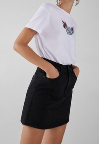White cotton t-shirt with colorful embroidered "ame" and wing design, paired with a black denim mini skirt featuring front pockets and a zipper.