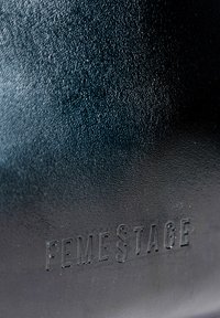 Borsa in pelle nera textured con logo "FEMESTAGE" in rilievo. La superficie presenta una finitura granulosa con leggero lucido.