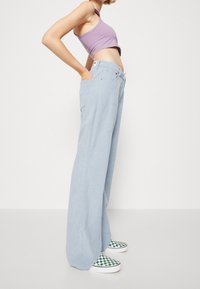 Person som bär ljusblå vida byxor, lila croppad topp och grön-vit rutiga slip-on skor, står med händerna i fickorna.