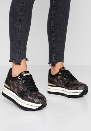 Nogi w czarnych dżinsach z postrzępionym brzegiem, noszące czarno-brązowe sneakersy na platformie ze złotymi akcentami i wzorzystym logo na szarej podłodze.