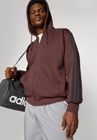 Bordeaux kleuren hoodie met ritssluiting en zwarte strepen op de mouwen, kangoeroe-zakken en verstelbare trekkoordcapuchon. Gepaard met lichtgrijze shorts en een zwarte tas.