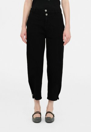 Zwarte, high-waist cropped broek met knoopdetails aan de zoom, gedragen met zwart-witte platte schoenen met patroon en een zwarte mouwloze top.