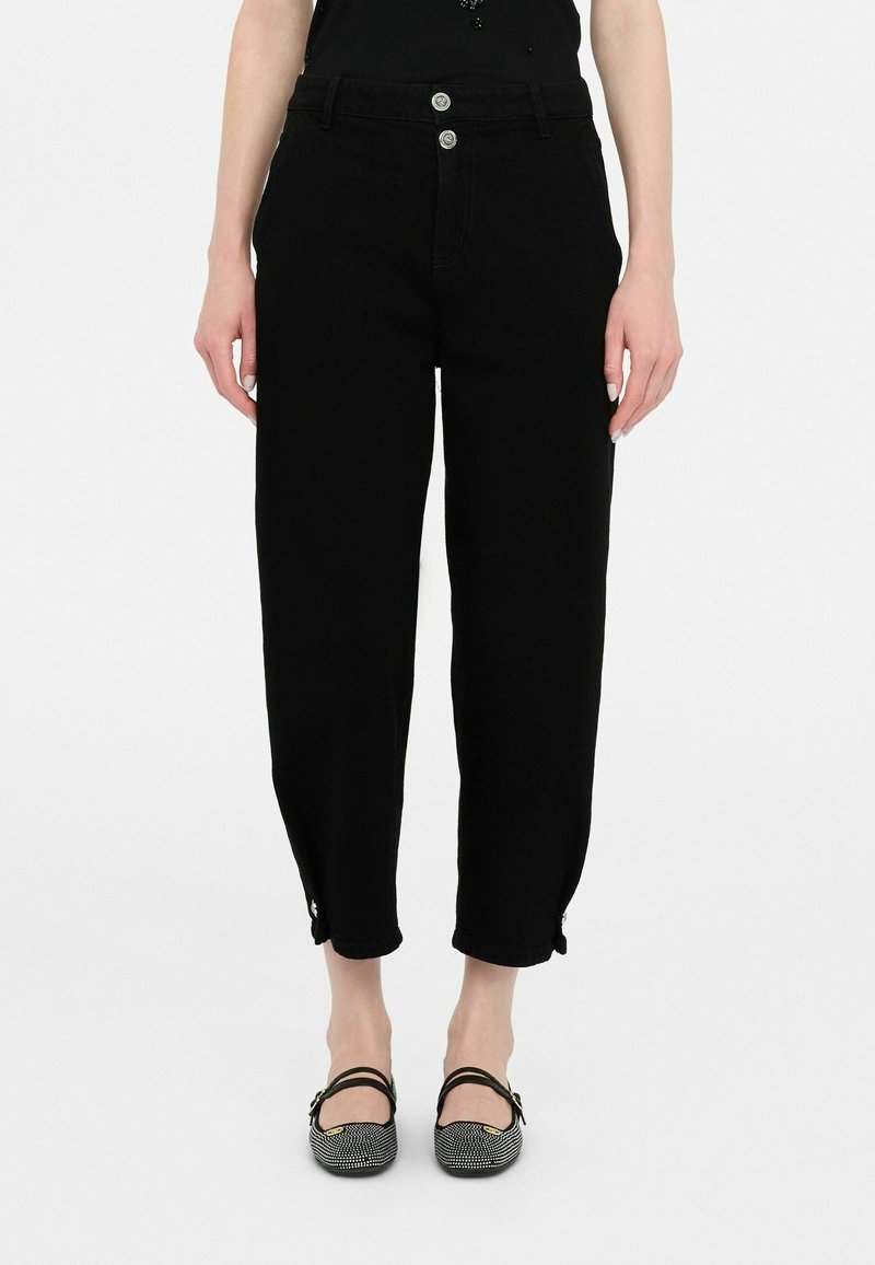 Pantalon court noir taille haute avec ourlets boutonnés, porté avec des chaussures plates noires et blanches à motif et un haut noir sans manches.