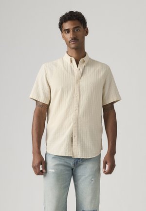 Homme aux cheveux courts et bouclés avec une moustache, portant une chemise beige à manches courtes boutonnée et un jean bleu clair déchiré, se tient devant un fond uni.