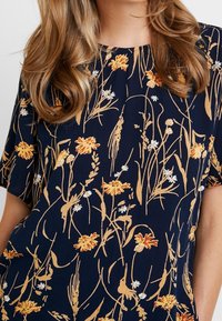 Mörkblå blus med korta ärmar, prydd med gula och vita blommönster samt en mjuk, texturerad tygkvalitet. Rund halsringning.
