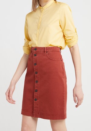 Gonna di jeans rossa con taglio dritto, con chiusura frontale a bottoni e due tasche laterali. Indossata con una camicia gialla con colletto.