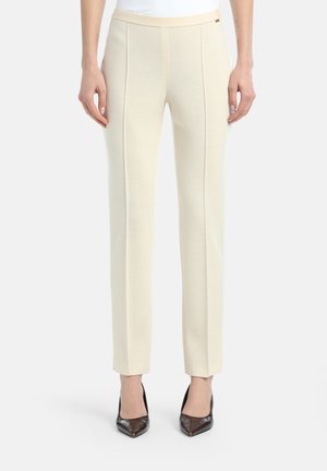 Pantalons de couleur crème avec une coupe slim, présentant une texture lisse, des coutures latérales et une coupe droite, associés à des chaussures à talons hauts sombres.