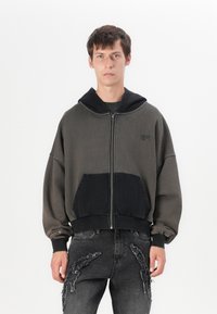 Fato de capuz oversized com fecho éclair em verde escuro e preto, apresentando um bolso em contraste, punhos canelados e uma textura suave.