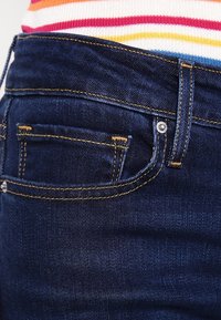 Mörkblå denimjeans med kontrasterande gula sömmar, försedda med en bakficka och metallknappdetalj längs midjan.