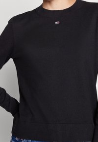 Suéter de punto negro con cuello redondo, puños y dobladillo acanalados, con un pequeño logo en el pecho. Textura suave, diseño ajustado.