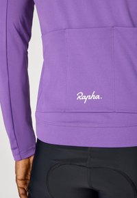 Ljubičasta majica dugih rukava za biciklističke sportove s glatkom teksturom, sa stražnjim džepom i bijelim logotipom "Rapha" blizu rubа. Crne biciklističke kratke hlače su vidljive ispod.