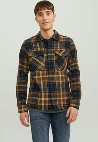 Camisa de cuadros en tonos marrón, azul marino y gris; presenta dos bolsillos en el pecho, cierre frontal con botones y un cuello con solapa. Textura de tela suave.