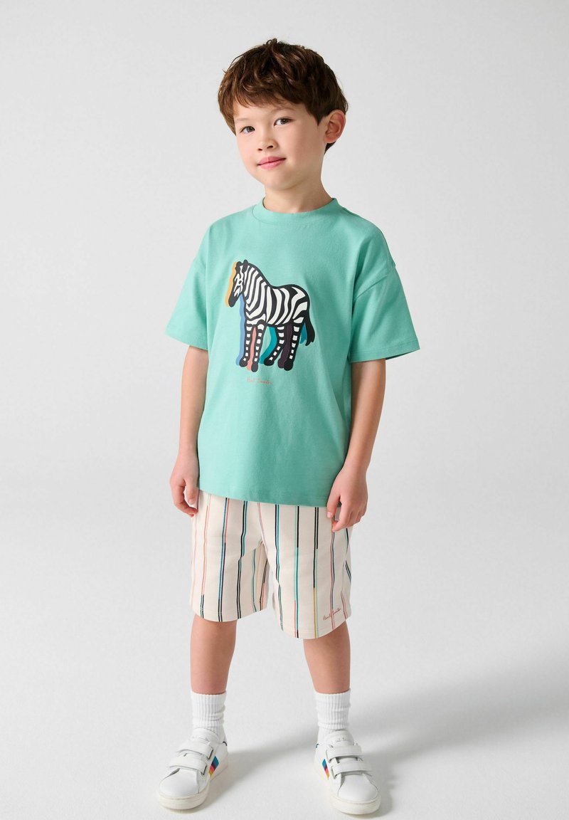 Ung dreng iført en turkis t-shirt med zebra-print, stribede shorts, hvide sokker og hvide sneakers med regnbuedetaljer.