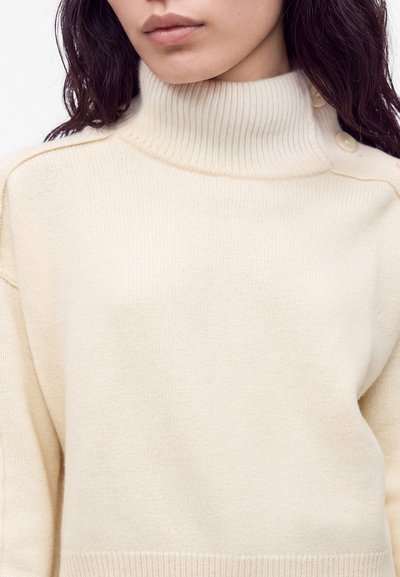 Femme portant un pull à col roulé côtelé couleur crème avec des boutons sur l'épaule droite et manches longues.