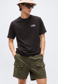 Άνδρας που φοράει μπεζ καπέλο κάδο, μαύρο t-shirt Patagonia, λαδί cargo σορτς και σκουρόχρωμα γυαλιά ηλίου, στέκεται με τα χέρια στις τσέπες.