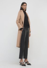 Cappotto di lana beige con design a bottoni singoli, indossato sopra una maglietta nera e pantaloni in pelle nera aderenti, abbinato a mule nere con tacco.