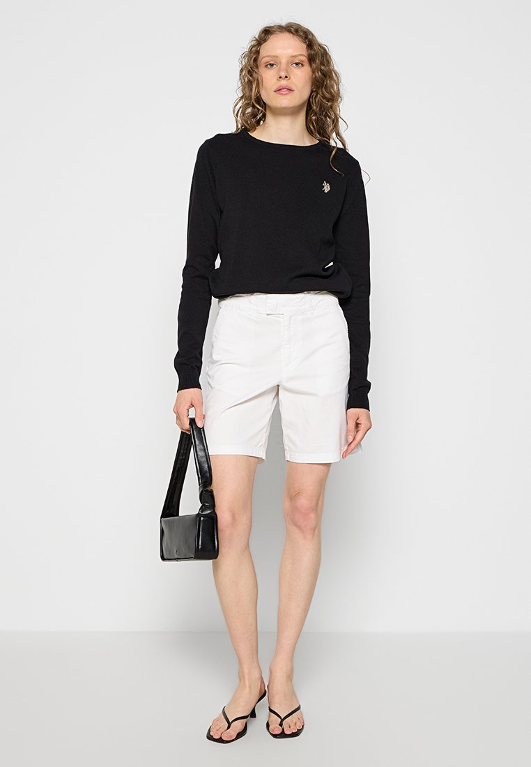 Schwarzer Pullover mit langen Ärmeln, weiße Shorts, schwarze Handtasche und schwarze Flip-Flops. Glatte Texturen mit einer taillierten Passform und subtilen Logodetails.