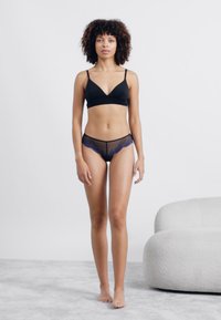 Čierny bralet s tenkými ramienkami v kombinácii s čiernymi priesvitnými šortkami na bokoch so zdobným modrým čipkovým lemovaním. Mäkké, štruktúrované pozadie.