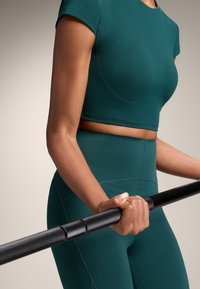 Zeleno moder športni crop top s kapicami, v kombinaciji z visokimi hlačnicami. Oseba drži črno vadbeno palico. Gladek, raztegljiv material.
