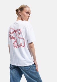 Hvid bomulds t-shirt med en rød broderet sløjfe og teksten "Make A Wish" på ryggen. Afslappet pasform med korte ærmer.