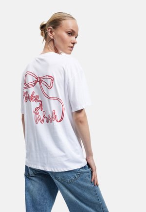 Witte katoenen t-shirt met een rode geborduurde strik en de tekst "Make A Wish" op de achterkant. Relaxed fit met korte mouwen.