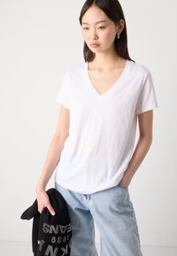 Vit bomulls V-ringad t-shirt med korta ärmar, med en subtil broderad logotyp på bröstet. Bärs tillsammans med ljusblå denimbyxor.