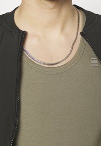 Chaqueta negra con cremallera y cuello de canalé sobre una camiseta de tirantes ajustada verde oliva. Collar de cadena plateada se encuentra por encima del escote.