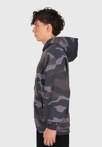 Ung mand i mørkegrå og sort camouflage-hoodie, stående i profil mod en ensartet lys baggrund.