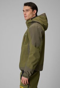 Jeune homme portant une veste d'extérieur à capuche verte et olive ainsi qu'un pantalon, debout de profil sur un fond gris uni.