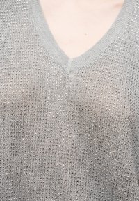 Femme portant un haut argenté à col en V recouvert de petits sequins métalliques sur la poitrine et les épaules.